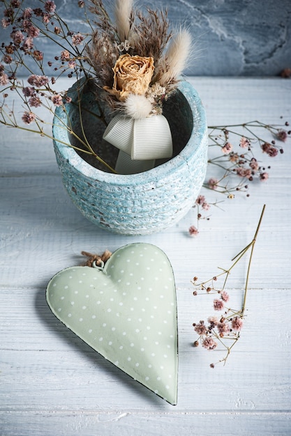 Ramo de flores secas en maceta azul con corazón verde y velas encendidas sobre mesa de madera blanca. Copiar espacio para texto, saludo, invitación