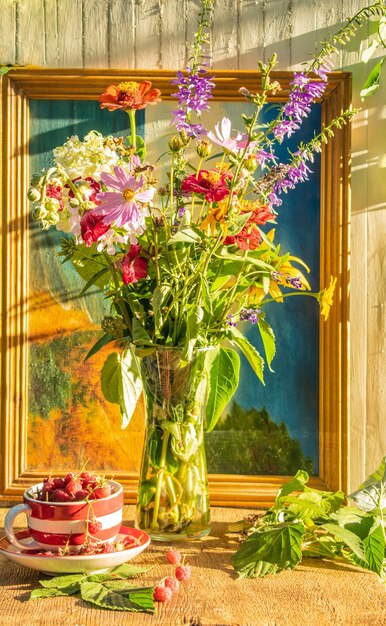 Un ramo de flores silvestres de verano en un jarrón de malla una taza con frambuesas sobre un fondo de madera