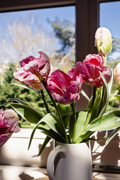 Ramo de flores de tulipán rosa con tallos verdes frescos en un jarrón de porcelana blanca en un marco de ventana blanco con fondo de cielo azul Idea de decoración floral de botánica colorida vibrante para el hogar