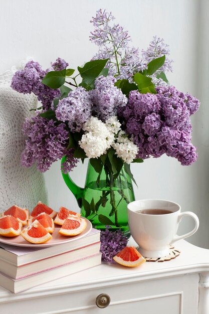 Un ramo fragante de lilas coloridas se encuentra en un jarrón de vidrio verde sobre un fondo blanco Junto a una taza de té caliente y pomelo en rodajas