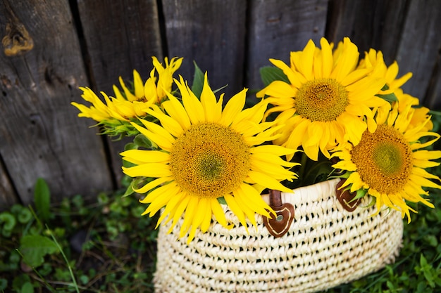 Un ramo de girasoles grandes en una bolsa de paja está de pie cerca de una vieja casa de madera.