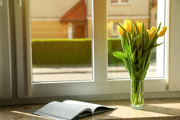 Ramo de hermosas flores de tulipán amarillo en jarrón de vidrio y libro abierto en el alféizar de la ventana Espacio para texto