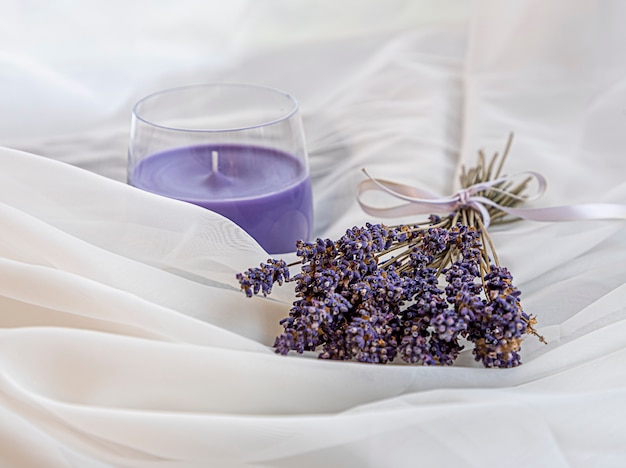 Ramo de lavanda seca atado con una cinta y la vela de lavanda de aroma descansa sobre una tela blanca y aireada. Enfoque selectivo con poca profundidad de campo. Armonía