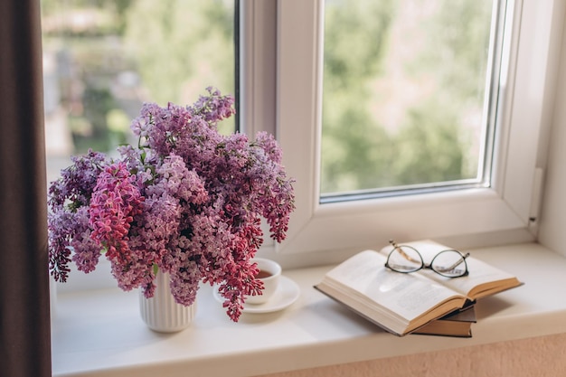 Ramo de lilas en una taza de café y libros en el alféizar de la ventana