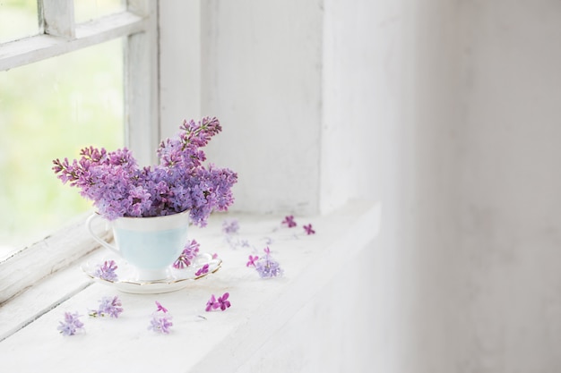 Ramo de lilas en taza de cerámica en el alféizar blanco viejo