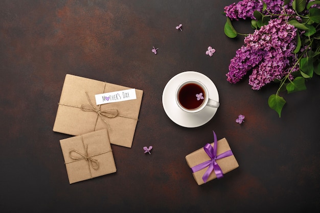 Un ramo de lilas con una taza de té, caja de regalo, sobre de artesanía, una nota de amor sobre fondo oxidado. Día de la Madre