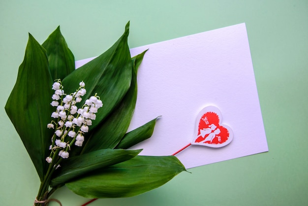 Un ramo de lirios del valle, una hoja de papel y un corazón en una pared verde.