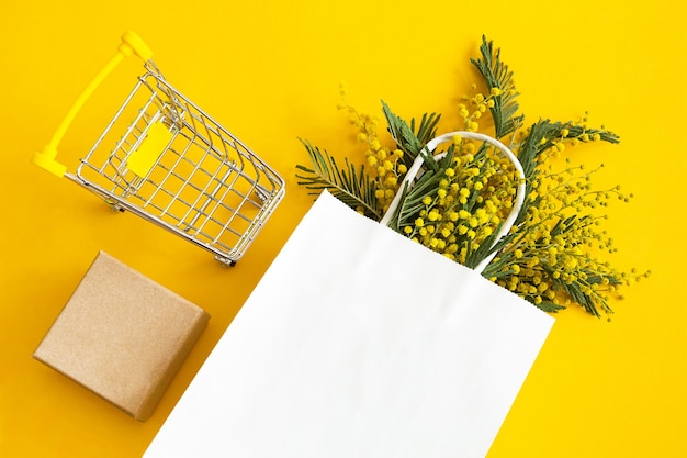 Un ramo de mimosa en una bolsa de papel blanca de imitación, una caja de regalo artesanal y un carrito de la compra.