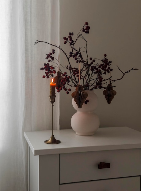 Ramo de Navidad casero con juguetes de artesanía de papel y una vela encendida en un candelabro de bronce en un cajón blanco