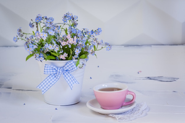 Ramo de nomeolvides en cubo de metal y una taza de té sobre fondo blanco.