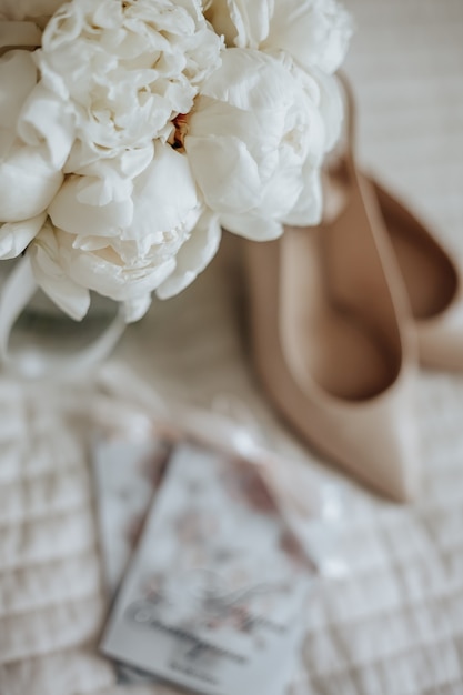 Ramo de novia de flores de peonías en un jarrón se encuentra en la cama de los recién casados con invitaciones y zapatos en el espacio de los vestidos de novia