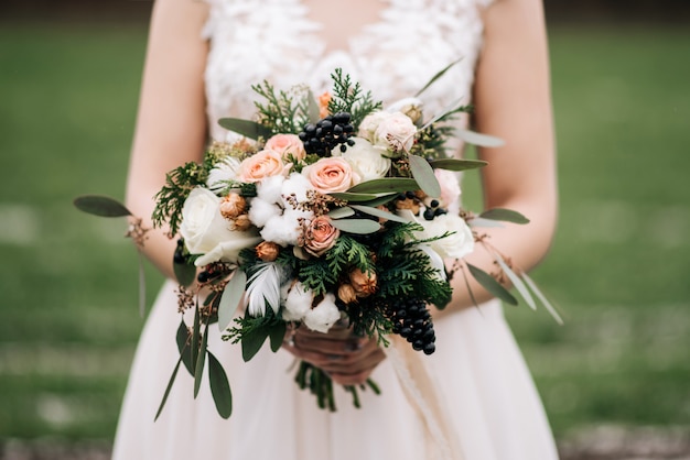 Ramo de novia de invierno con rosas, algodón, abeto, plumas, flores secas en las manos de la novia.