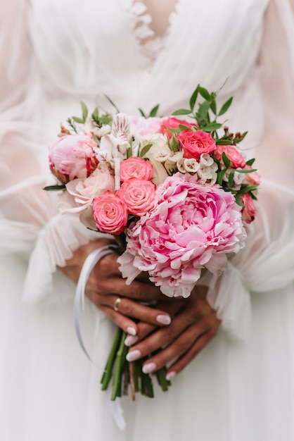 Ramo de novia de rosas blancas y rosadas y peonías en manos de la novia en el fondo de un vestido blanco