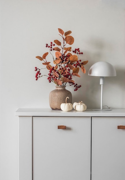 Ramo de otoño en un jarrón de cerámica, lámpara de mesa de estilo escandinavo, calabazas decorativas sobre una cómoda blanca