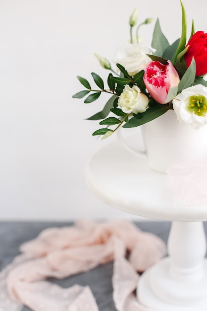 Ramo de primavera en florero blanco sobre soporte blanco de madera. Rosas, tulipanes y lisianthus.