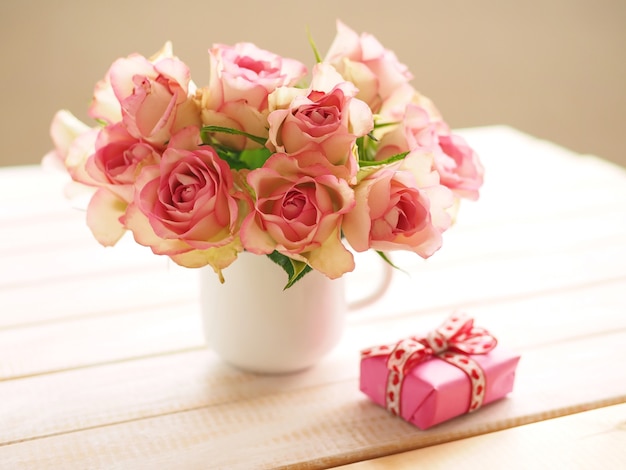 Ramo de rosas rosadas en taza blanca sobre mesa de madera y caja de regalo