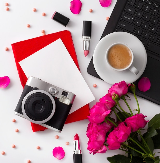 Ramo de rosas, una taza de café, una computadora portátil, una cámara, una libreta y un lápiz labial en la tabla blanca.
