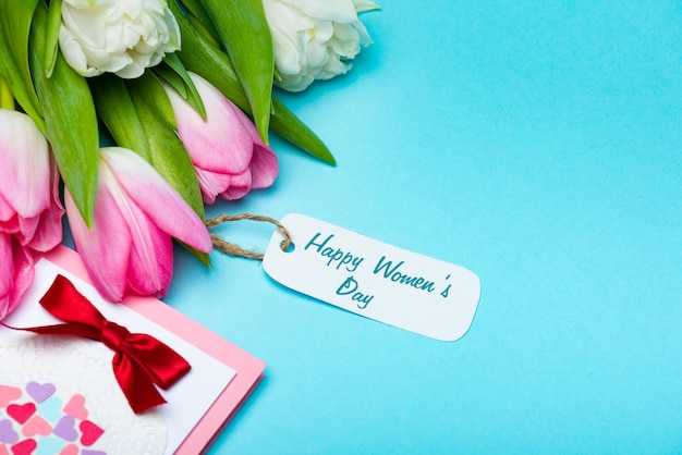 Ramo de tulipanes con letras de feliz día de la mujer en la etiqueta de papel cerca de la tarjeta de felicitación en azul