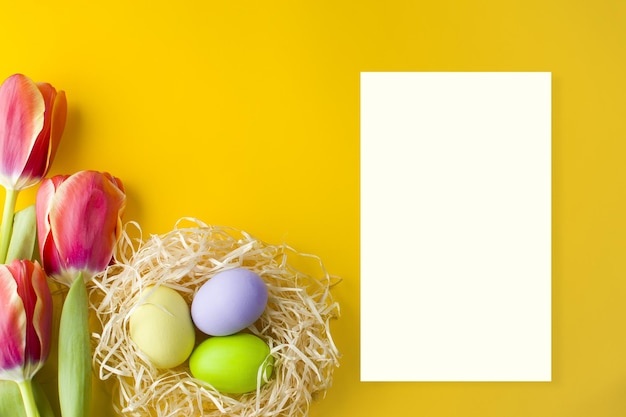 Un ramo de tulipanes, un nido con huevos de Pascua y una hoja de papel blanca sobre un fondo amarillo