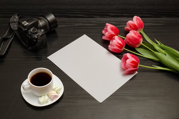 Ramo de tulipanes rosados, una taza de café, cámara réflex digital y una hoja de papel sobre una madera negra