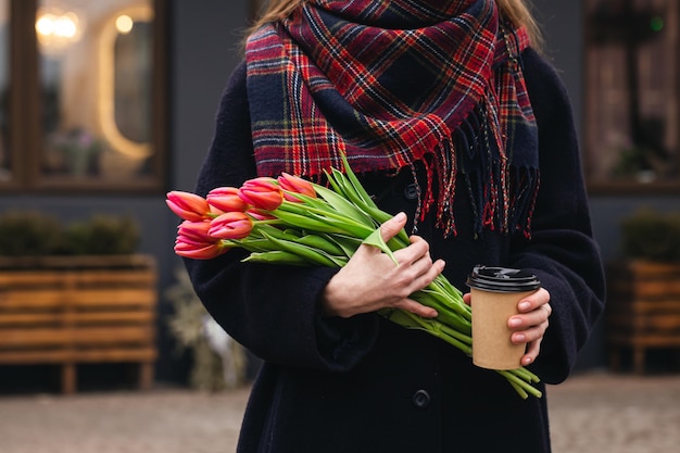 Un ramo de tulipanes rosados y un vaso de café de papel en manos femeninas afuera