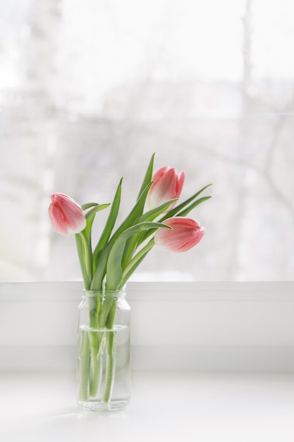 Foto un ramo de tulipanes rosas en un frasco de vidrio está en el alféizar de la ventana de la casa.