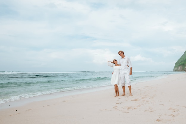 Rapariga e homem com roupas brancas, caminhando na praia branca em Bali e de mãos dadas