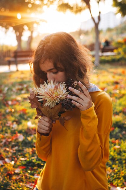 Foto rapariga encaracolada de suéter amarelo na grama com um buquê de folhas secas e flores de outono