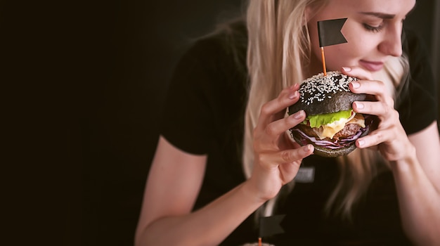 Rapariga loira com uma t-shirt preta com um grande hambúrguer preto nas mãos