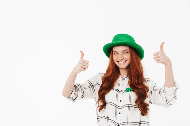 Rapariga ruiva feliz com chapéu verde, comemorando o Dia de São Patrício isolado sobre uma parede branca, com o polegar para cima