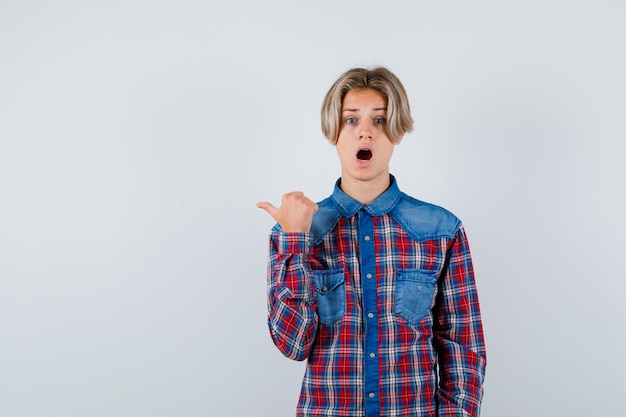 Rapaz adolescente apontando para o lado esquerdo com o polegar em uma camisa quadriculada e olhando perplexo, vista frontal.