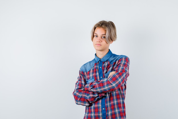 Rapaz adolescente em pé com os braços cruzados na camisa quadriculada e parecendo confiante, vista frontal.