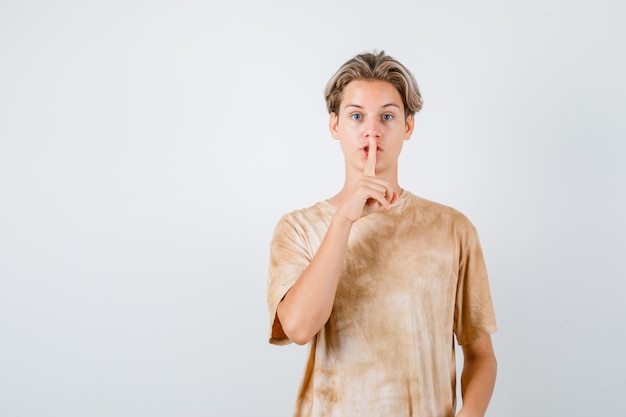 Rapaz adolescente em t-shirt mostrando gesto de silêncio e olhando cuidadoso, vista frontal.