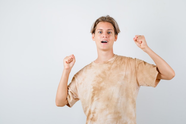 Rapaz adolescente em t-shirt mostrando o gesto do vencedor e olhando vitorioso, vista frontal.