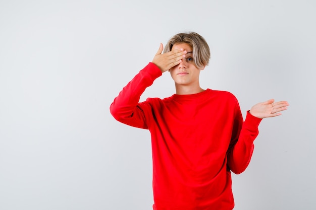 Rapaz adolescente fechando os olhos com a mão, mostrando um gesto de boas-vindas no suéter vermelho e olhando com medo, vista frontal.