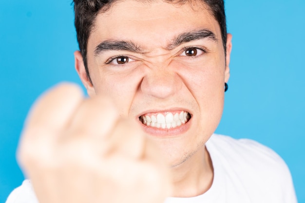 Rapaz adolescente hispânico irritado punho ameaçador e olhando para câmera isolada em fundo azul