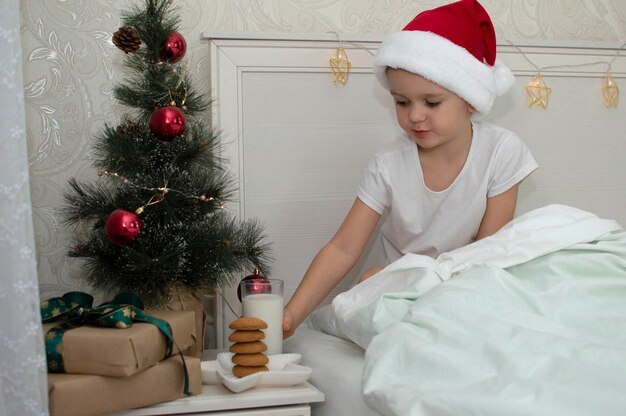 Rapaz adorável cozinha leite e biscoitos para o Papai Noel, espaço livre. Uma delícia tradicional é um biscoito de Natal e um copo de leite na mesinha de cabeceira perto da cama. O conceito de ano novo. Parentes