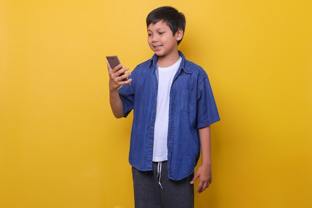 Rapaz asiático em estilo casual denim está sorrindo enquanto segura smartphone isolado em fundo amarelo O