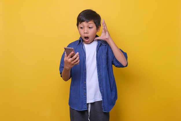 Rapaz asiático no estilo jeans casual está segurando o telefone e mostrando uma expressão surpresa isolada em amarelo