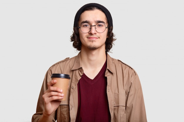 Rapaz bonito tem cabelo encaracolado, barba usa óculos redondos transparentes, mantém o café para ir, tem pausa, comunica-se com os amigos, modelos contra parede branca. Pessoas, estilo de vida, conceito de bebida