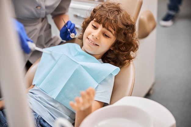 Rapaz cacheado de cabelo castanho sentado na cadeira de dentista a preparar-se para a consulta dentária