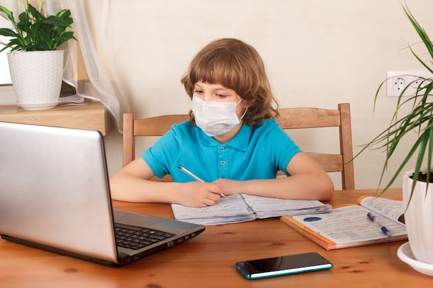 Rapaz com máscara médica no rosto, fazendo lição de casa e assistindo webinar no laptop. Educação a distância, ensino em casa, E-learning em casa durante o conceito de quarentena