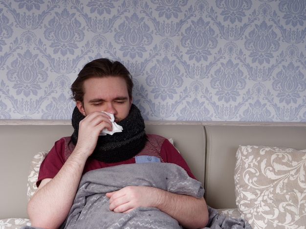 Rapaz de camiseta vermelha senta na cama e cuida do nariz doendo, ele está com o nariz escorrendo, o conceito de doença em casa