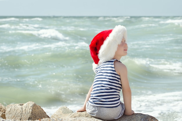 Rapaz de chapéu de Papai Noel e camiseta listrada, sentado na praia