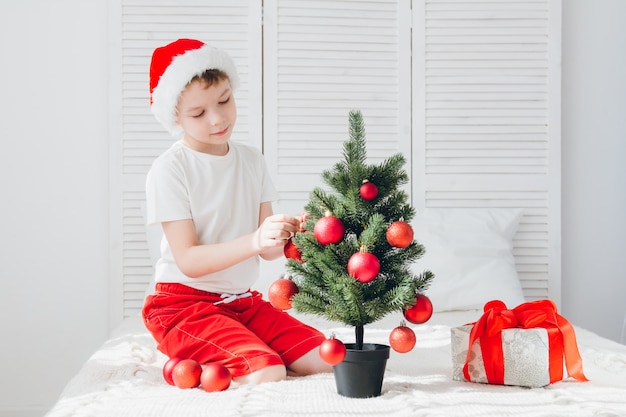 Rapaz de chapéu de papai noel vermelho decora uma pequena árvore de natal