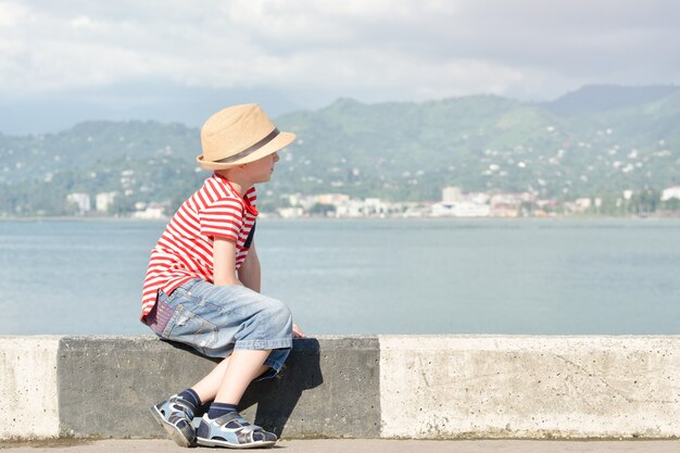 Rapaz de chapéu e camiseta listrada, sentado na praia e olha para o mar. vista lateral