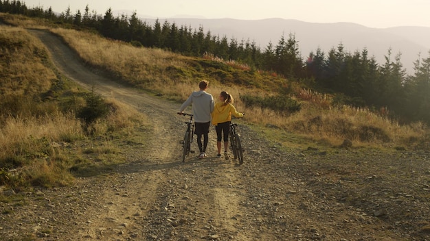 Rapaz e moça andando com bicicletas nas montanhas Ciclistas olhando a paisagem de verão Homem ruivo apontando com o dedo Casal aproveitando o fim de semana juntos