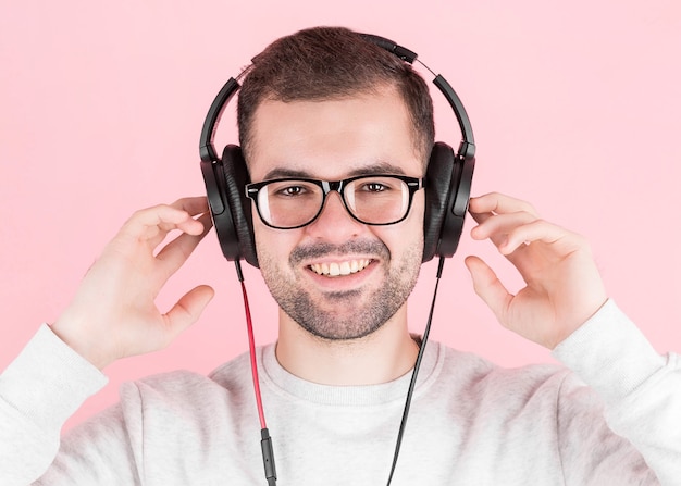 Rapaz feliz e fofo ouve música em grandes fones de ouvido brancos sobre um fundo rosa, segura-os, em um moletom branco, com um sorriso