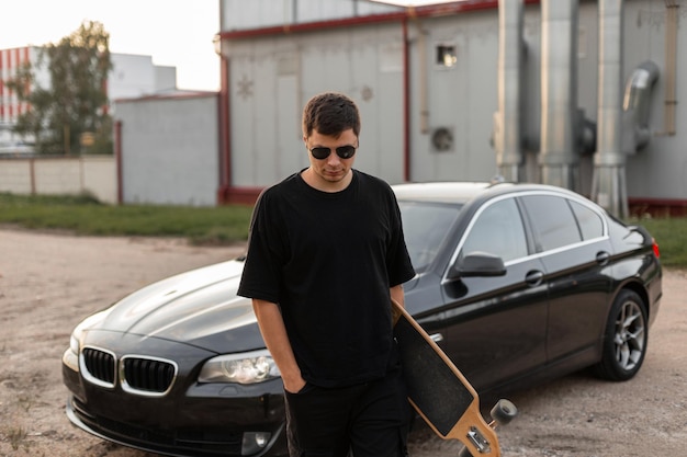 Rapaz hipster com óculos escuros em roupas pretas com longboard caminha ao ar livre perto de um carro e prédio