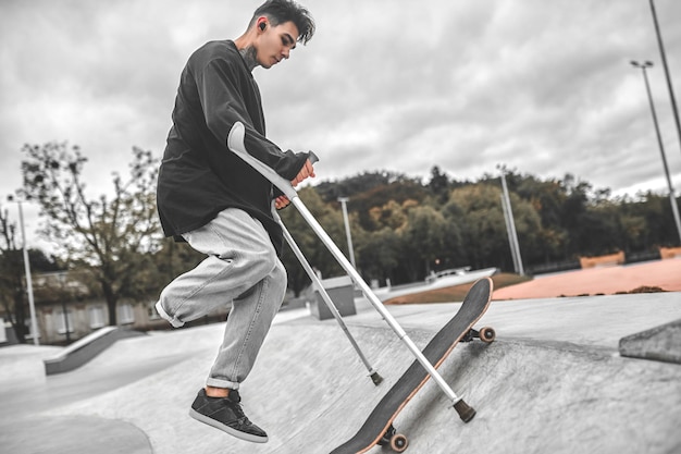 Rapaz pendurado sobre o skate e apoiado na rampa com muletas